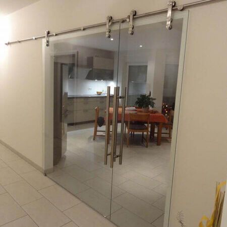 Vor der Wand laufende, asymmetrisch geteilte, doppelflügelige Glasschiebetür mit sichtbarem Design-Rollensystem